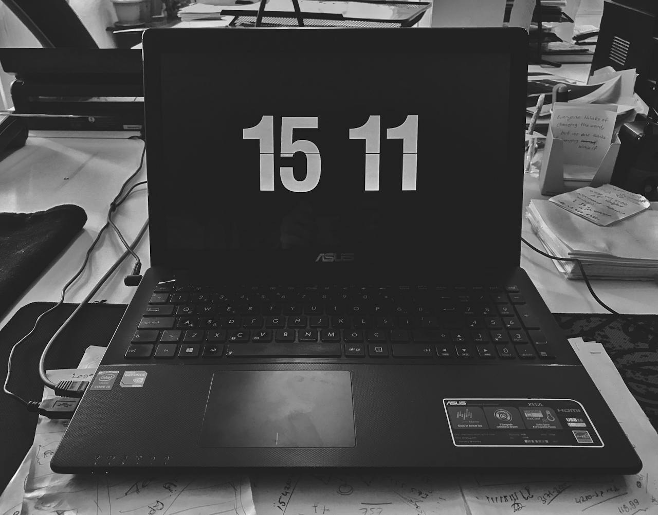 Black and white photo of a laptop displaying time, surrounded by desk items in a workspace.